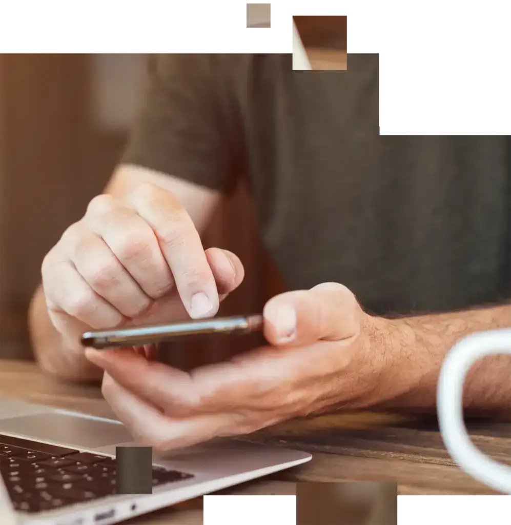 Mann tippt mit einem Finger auf ein Smartphone, vor ihm liegt ein Laptop auf einem Holztisch