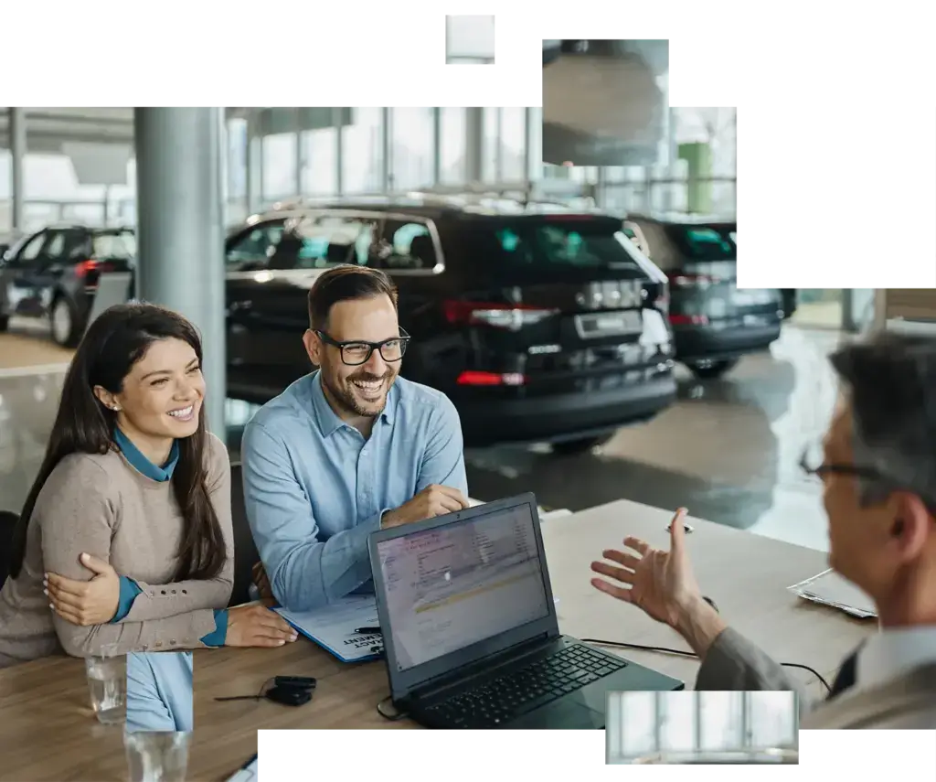 Paar sitzt lächelnd an einem Tisch im Autohaus und spricht mit einem Berater, auf dem Tisch steht ein Laptop