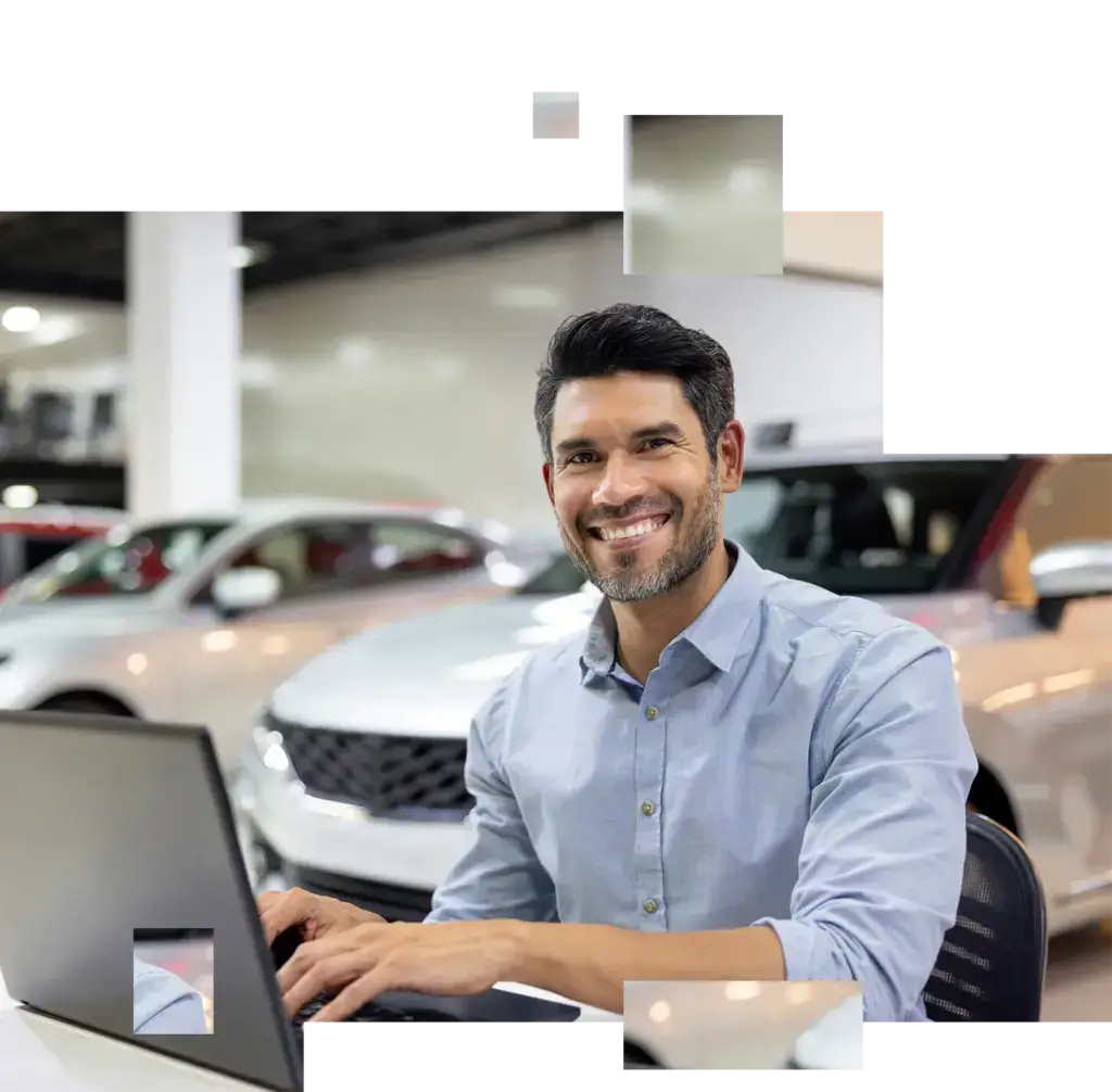 Mann mit Hemd sitzt lächelnd vor Laptop in einem Autohaus mit mehreren Fahrzeugen im Hintergrund